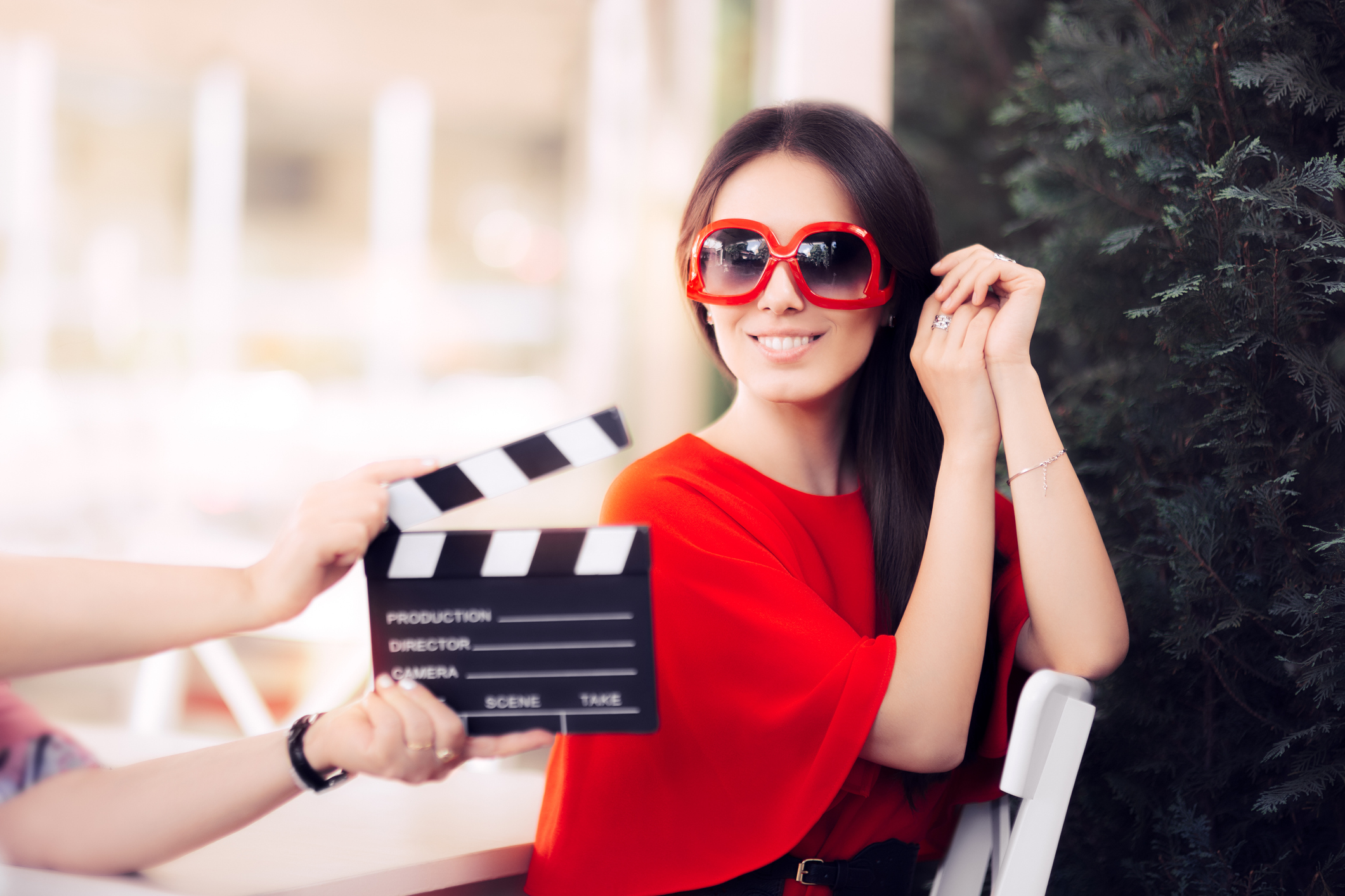 An actress in a red dress and oversized glasses smiles behind the film crew's clapper.