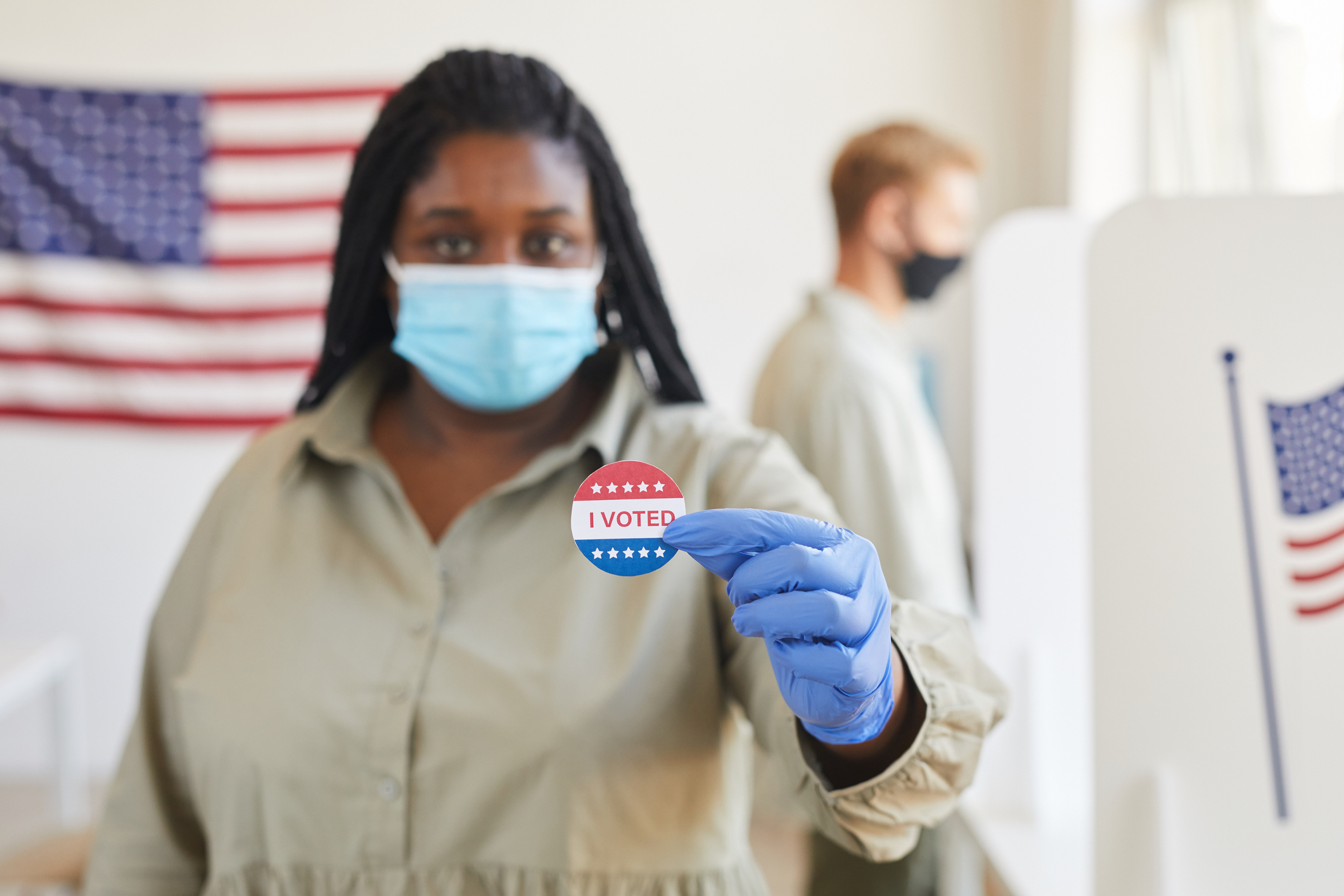 A masked poll worker presents an I VOTED sticker at a voting precinct.