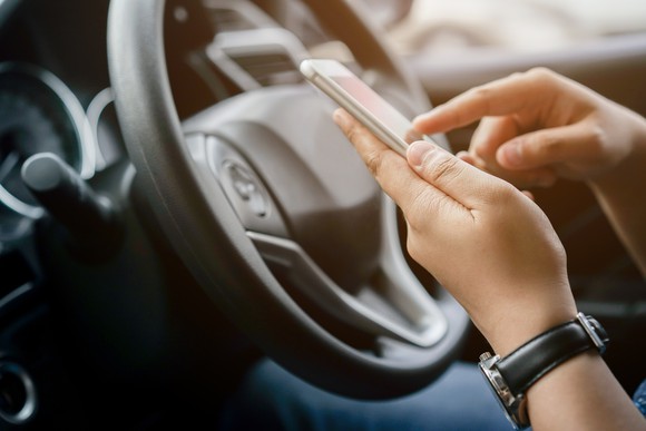 A pair of hands holds a cell phone in front of a steering wheel.