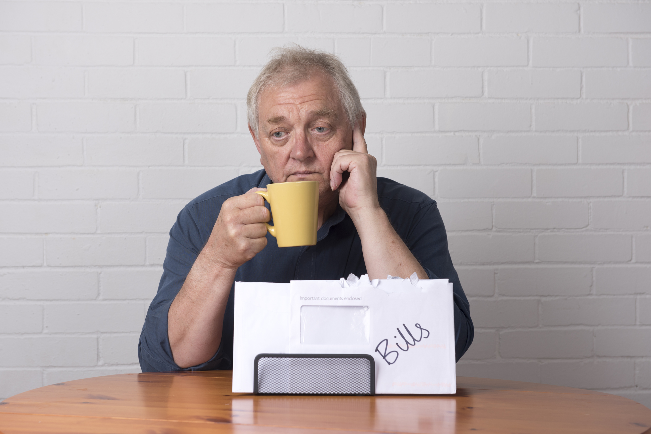 A visibly concerned senior man drinking from a coffee mug in front of a pile of bills.