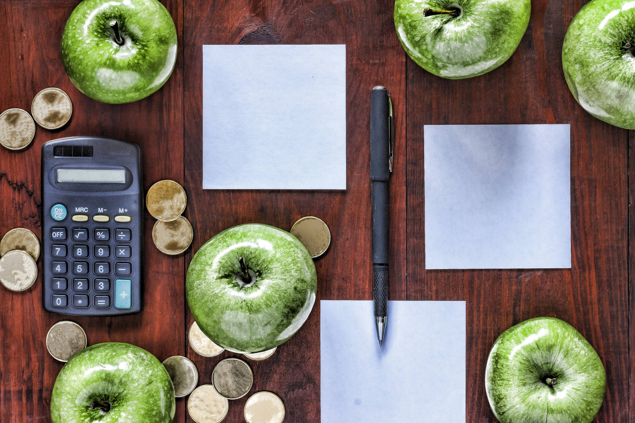 A desk with neatly arranged calculator, pen, papers, gold coins, and green apples.