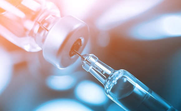 A glass vial with vaccine and syringe poking through stopper on top of vial.