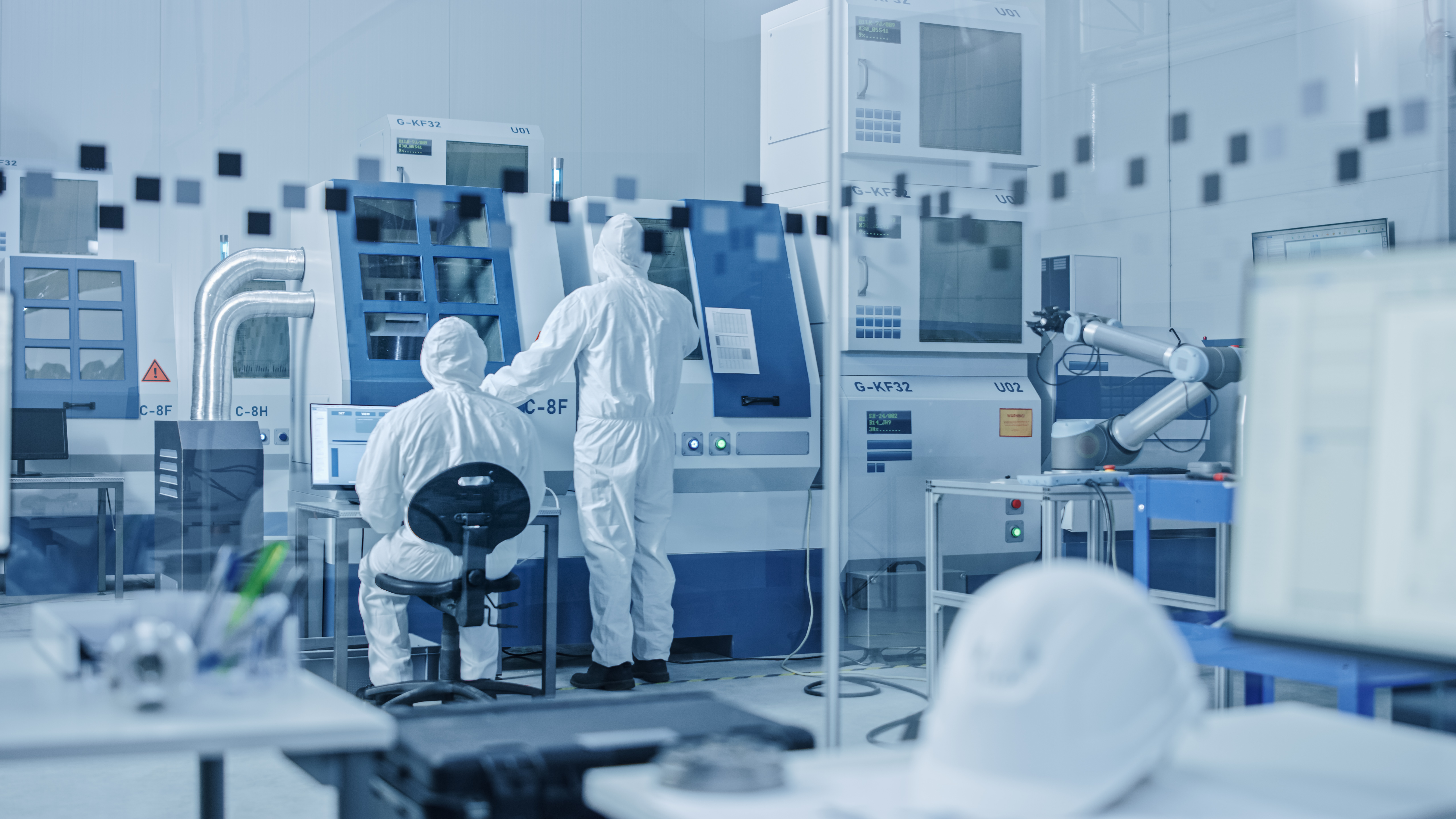 Two lab reachers dressed in protective gear face a large machine in a pharmaceutical lab colored mostly blue and white.