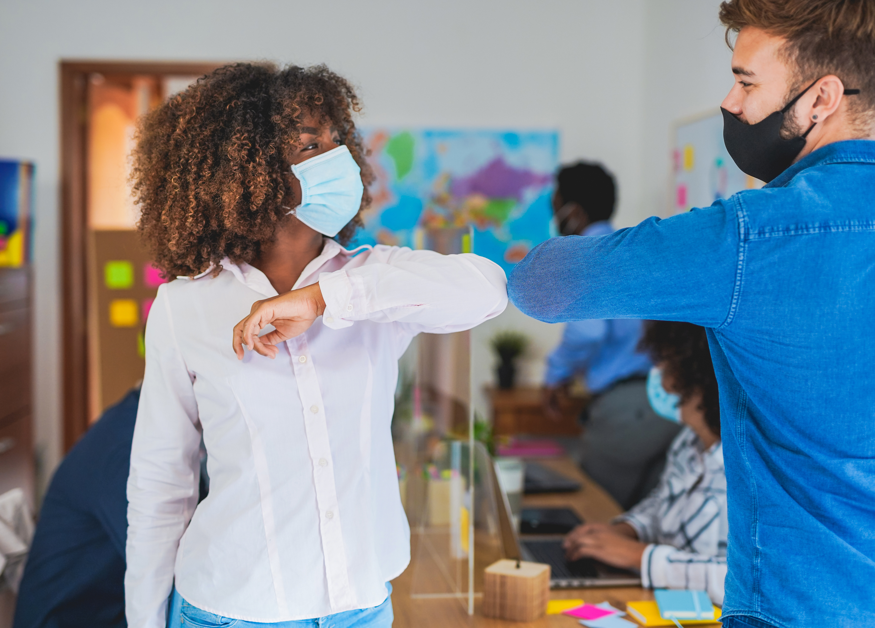 Two young people, both wearing masks, bump elbows in a busy office.