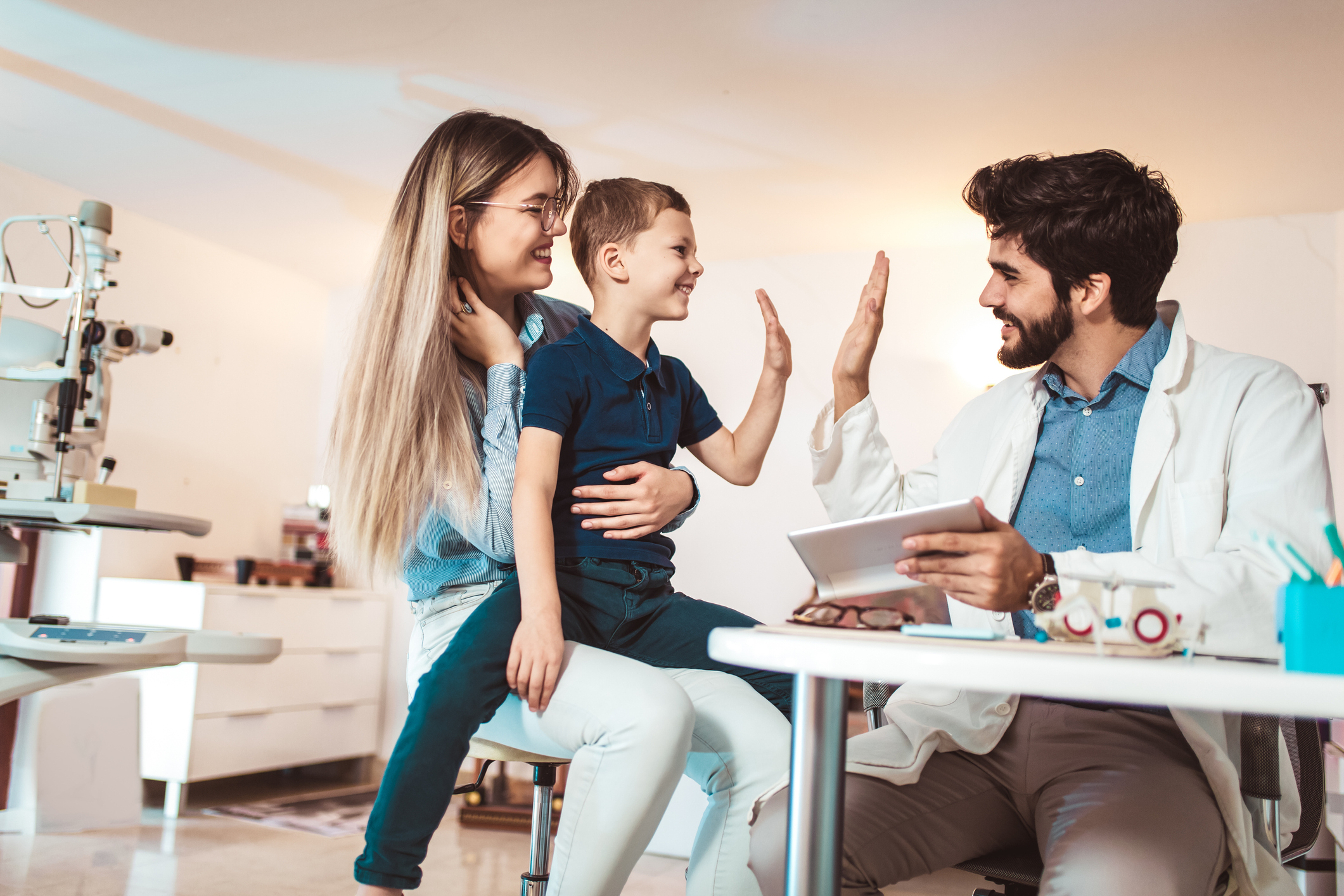 Healthcare provider with beard giving a high-five to a pediatric patient sitting on his mother's lap.