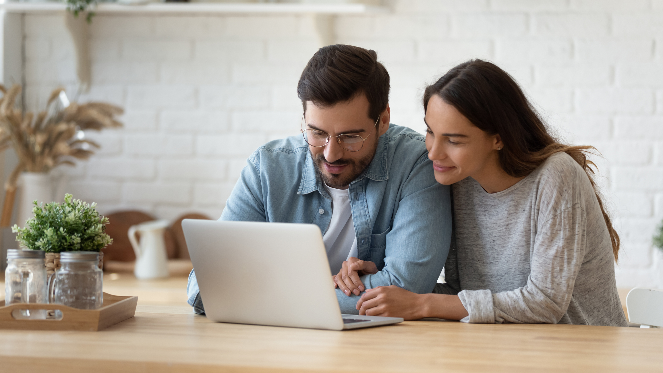 Sitting at a kitchen table, a man and woman look at a laptop.