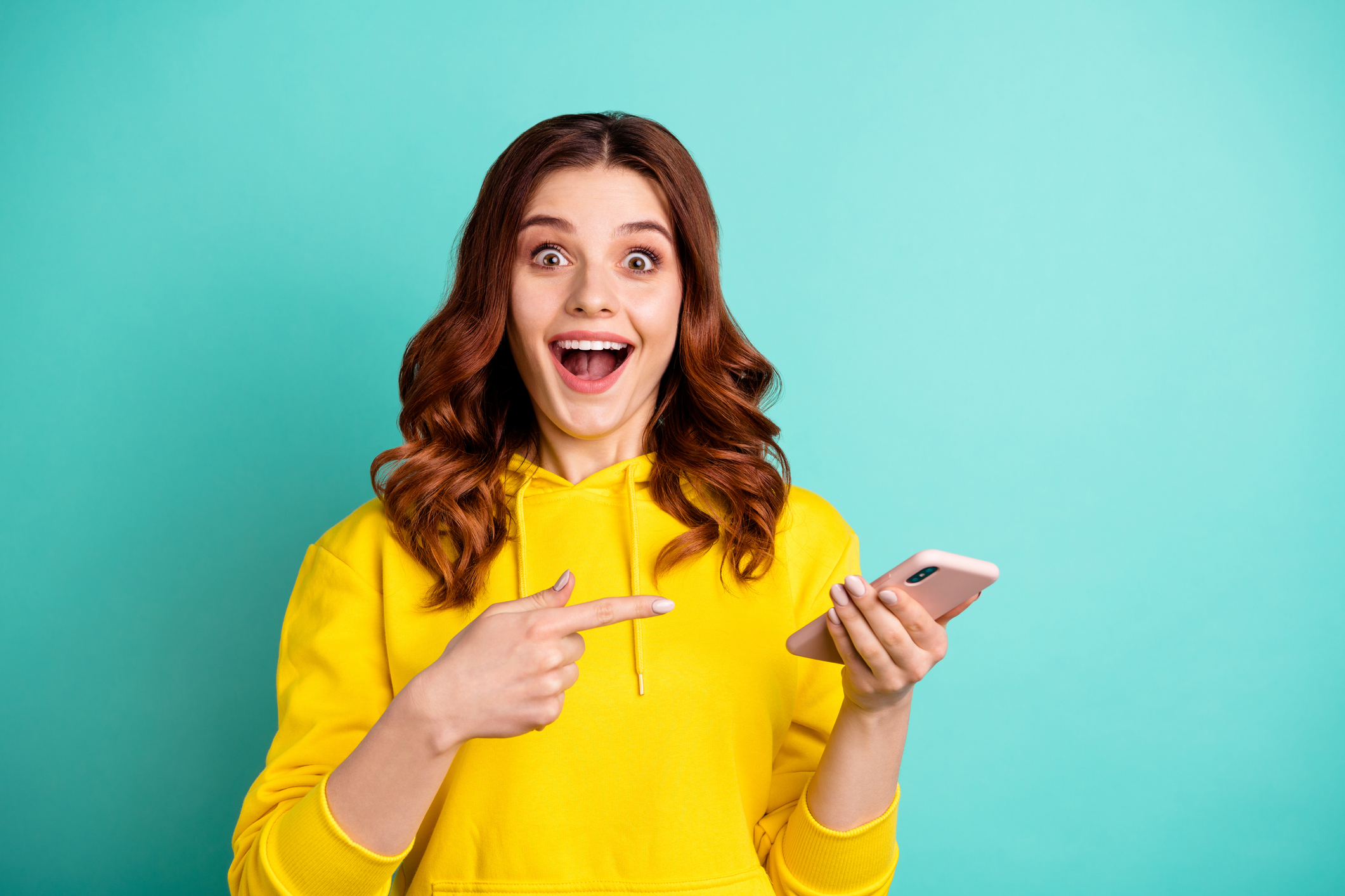 surprised young woman holding phone in yellow sweatshirt against light blue background