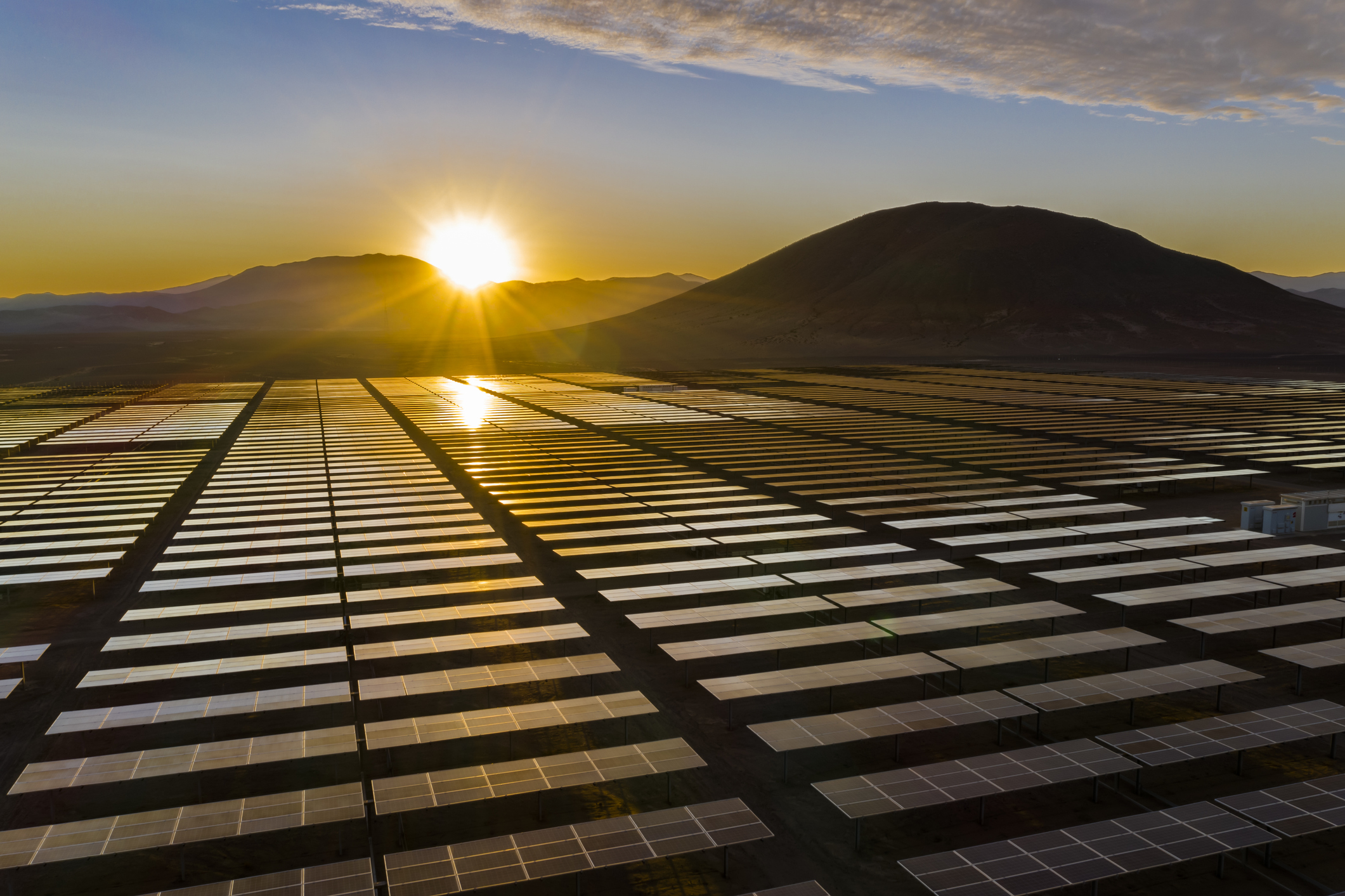 A sun rises over a mountain down on a field of solar panels.