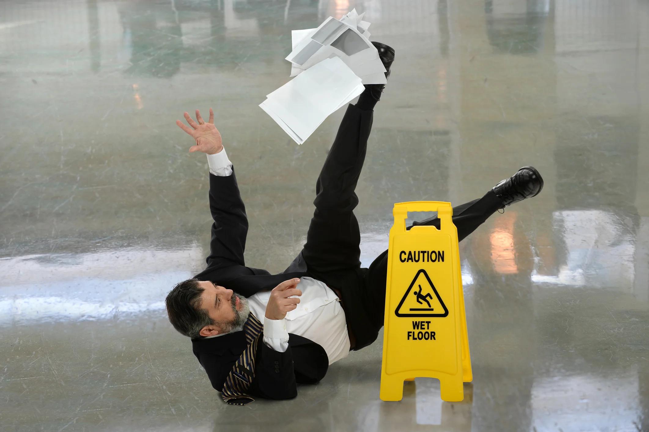 Man in suit falling down and spilling papers next to caution sign saying Wet Floor