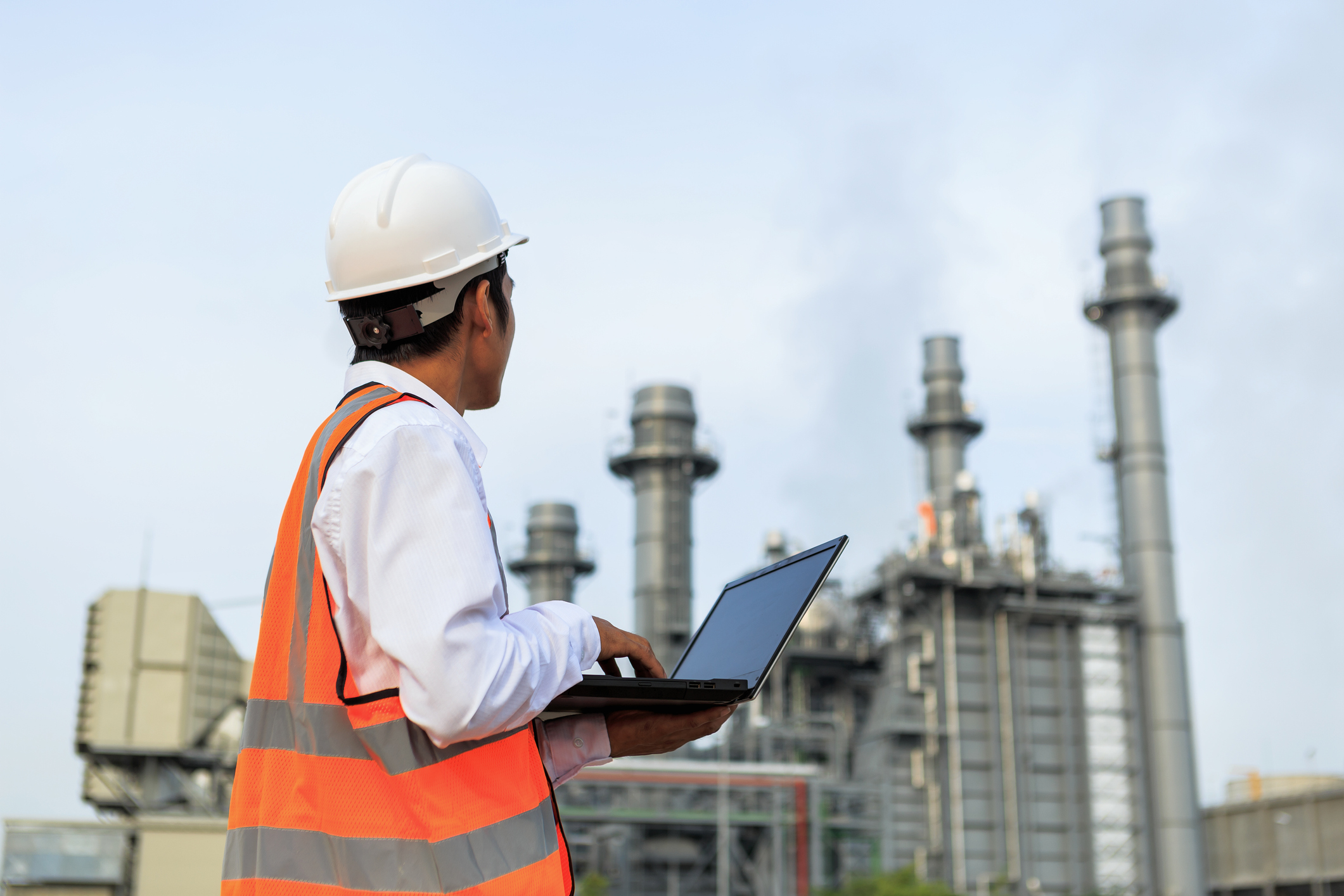 An engineer surveying an oil and gas refinery while holding an open laptop. 