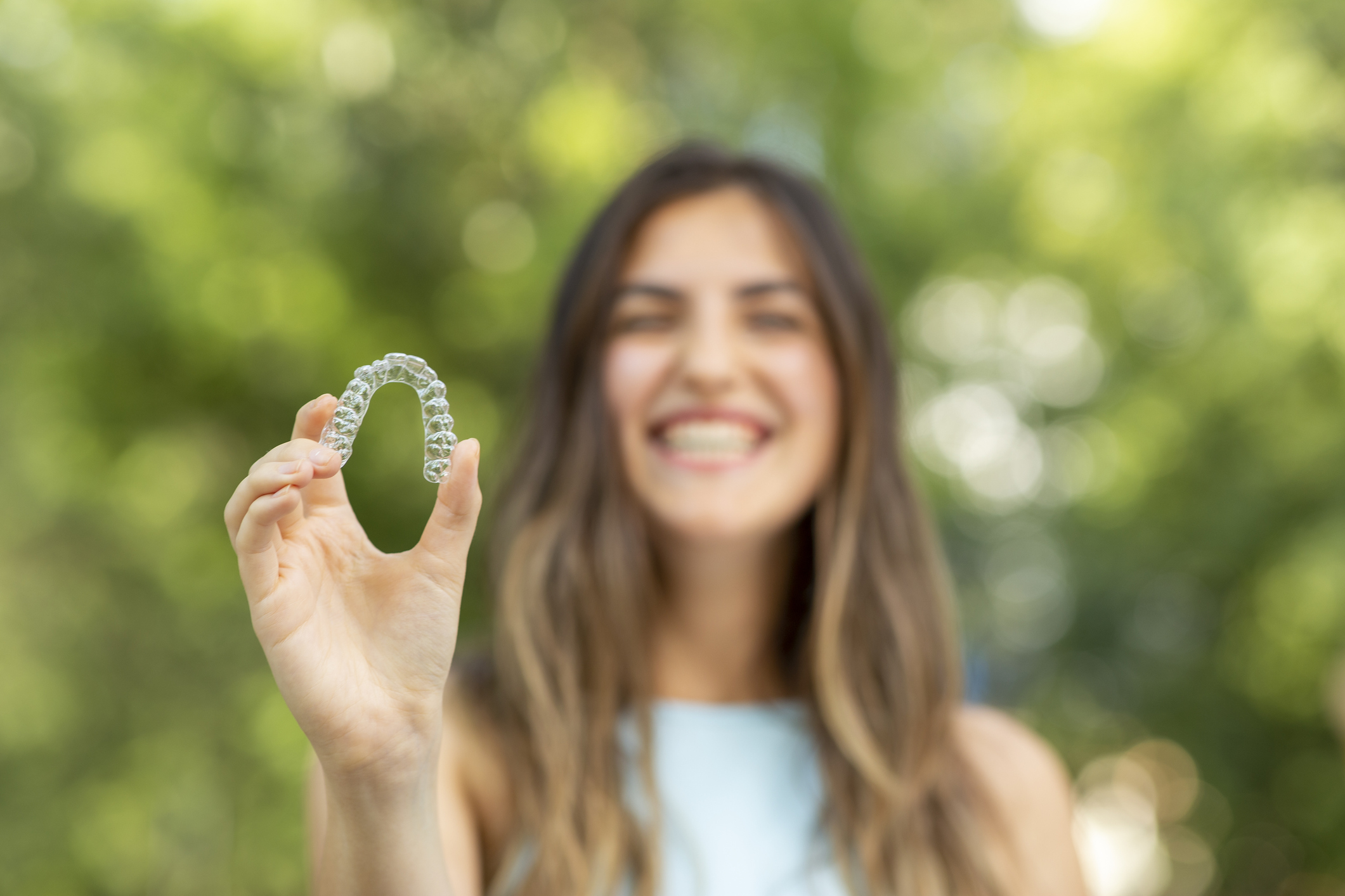 Smiling woman blurred in background holding up a clear invisaligner.