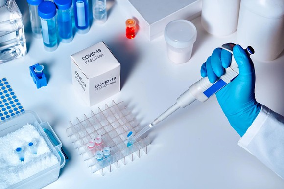 Hand with blue glove holding dropper putting blue liquid into tubes, with box labeled COVID-19 on lab table with other supplies.
