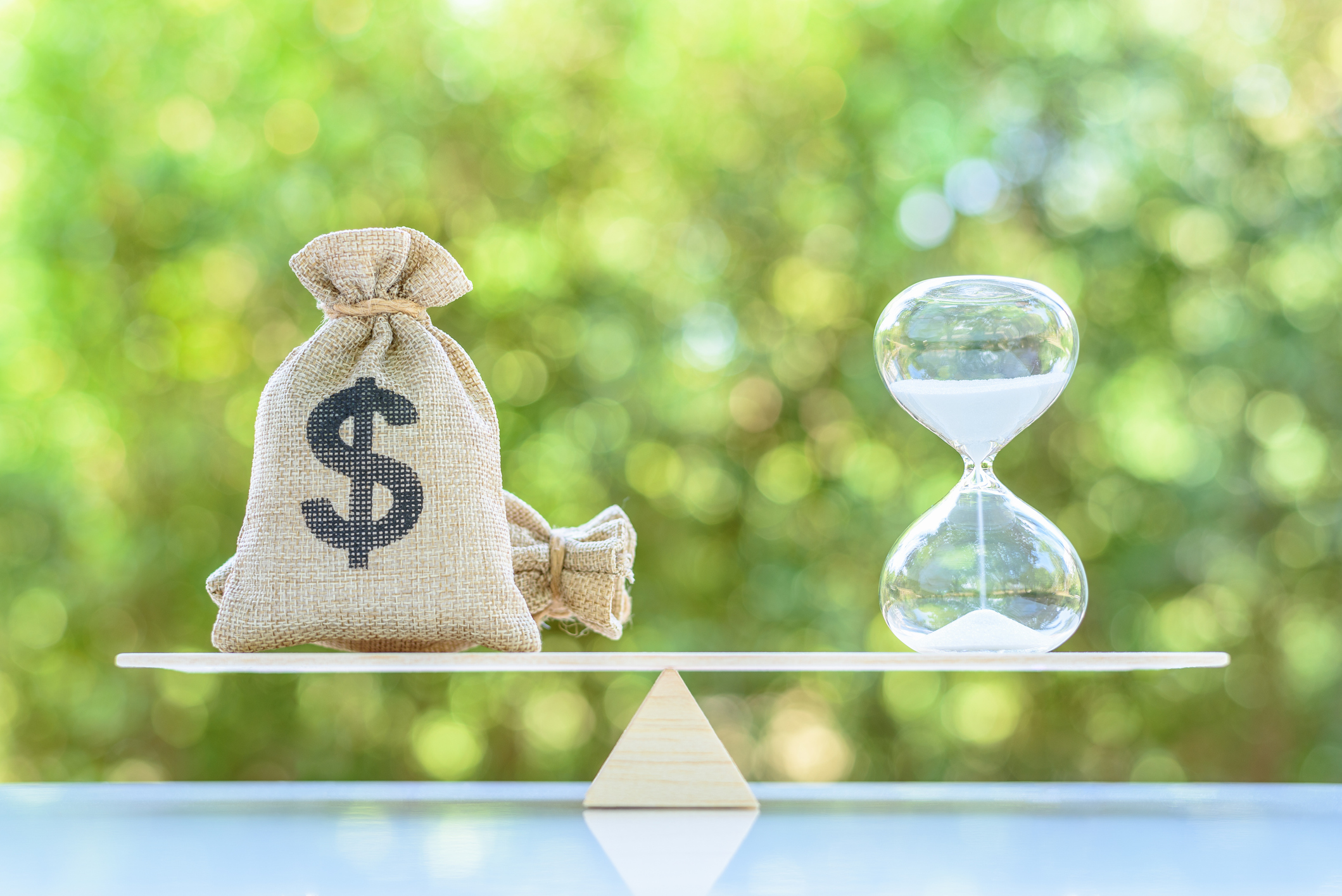 A bag with a money symbol is weighed against an hourglass on a scale.
