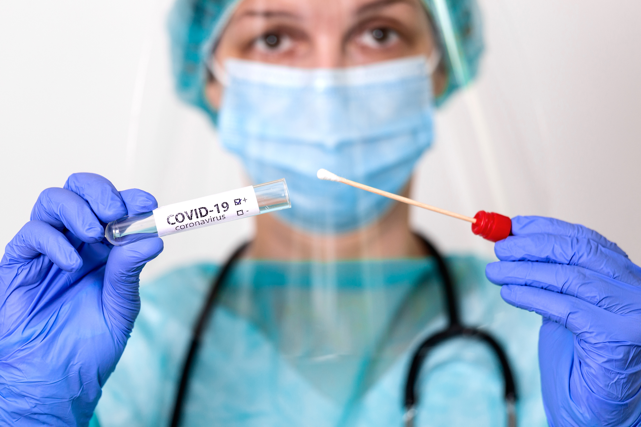 Medical profession in teal gown and full personal protective equipment holds up a COVID-19 test swab.
