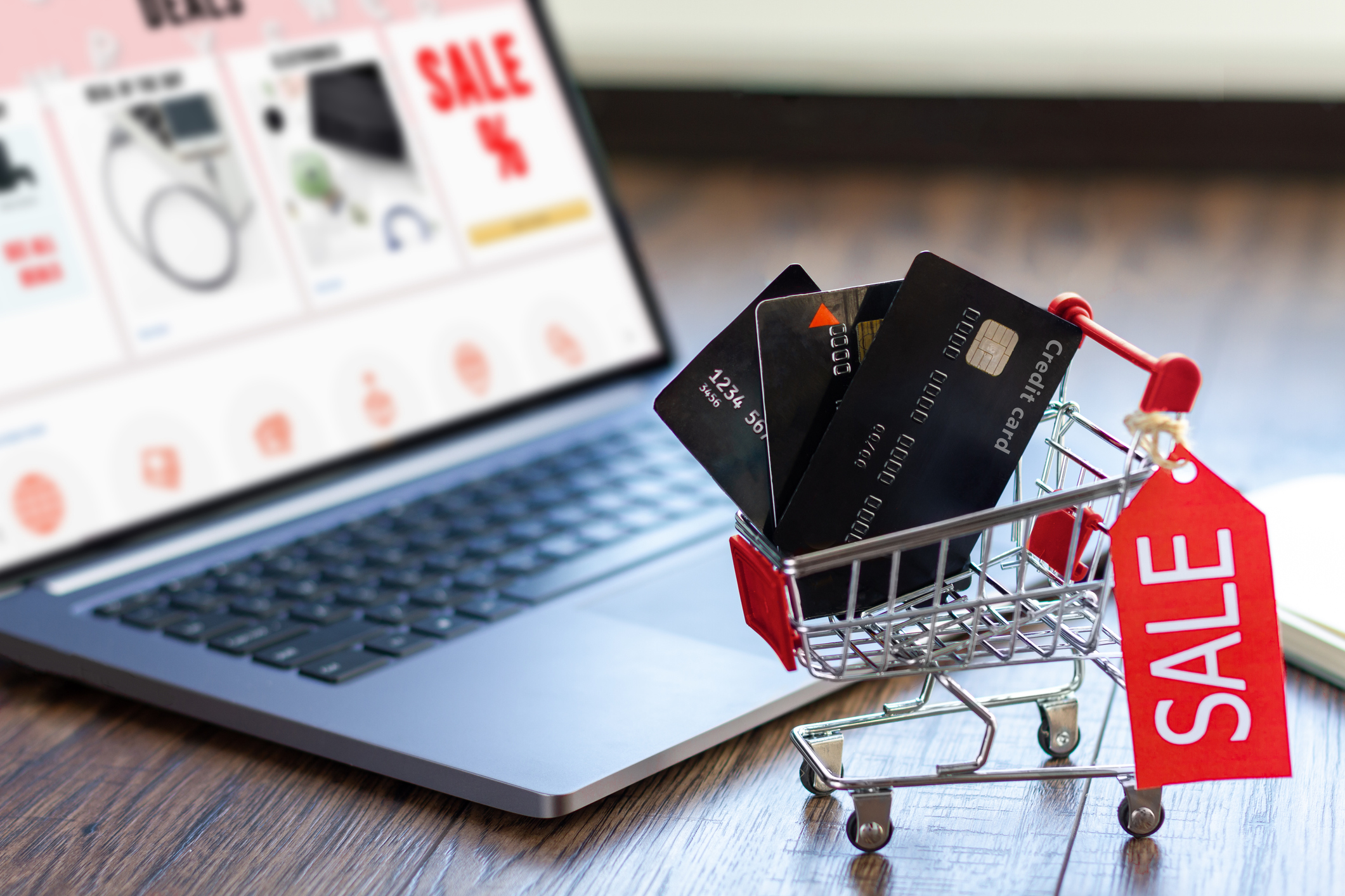 A laptop displaying a shopping sale. A toy shopping cart with a sale tag filled with credit cards.