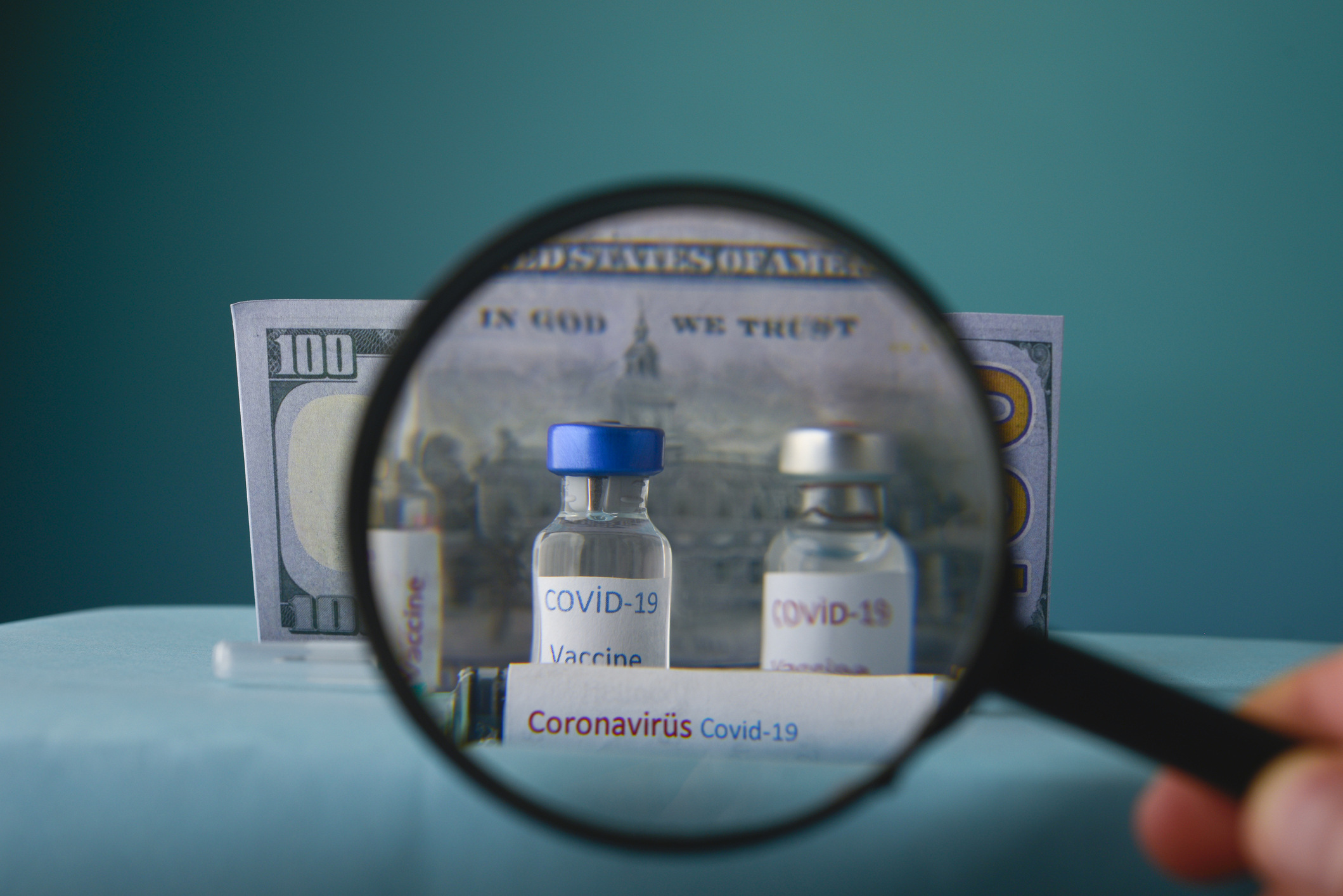 A magnifying glass held in front of two COVID-19 vaccine vials with a $100 bill behind them