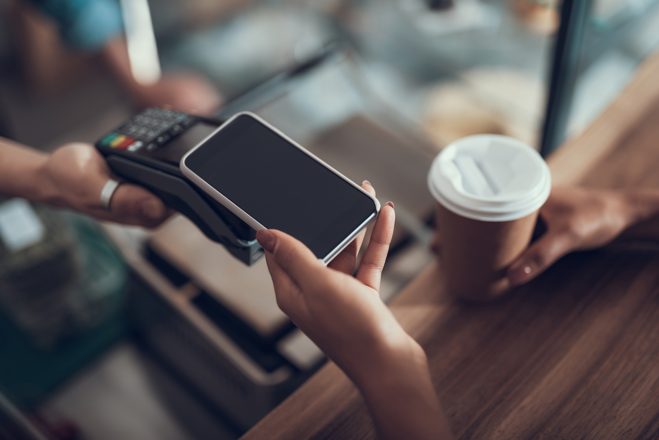 A hand holding a smartphone up to a wireless card machine at checkout.
