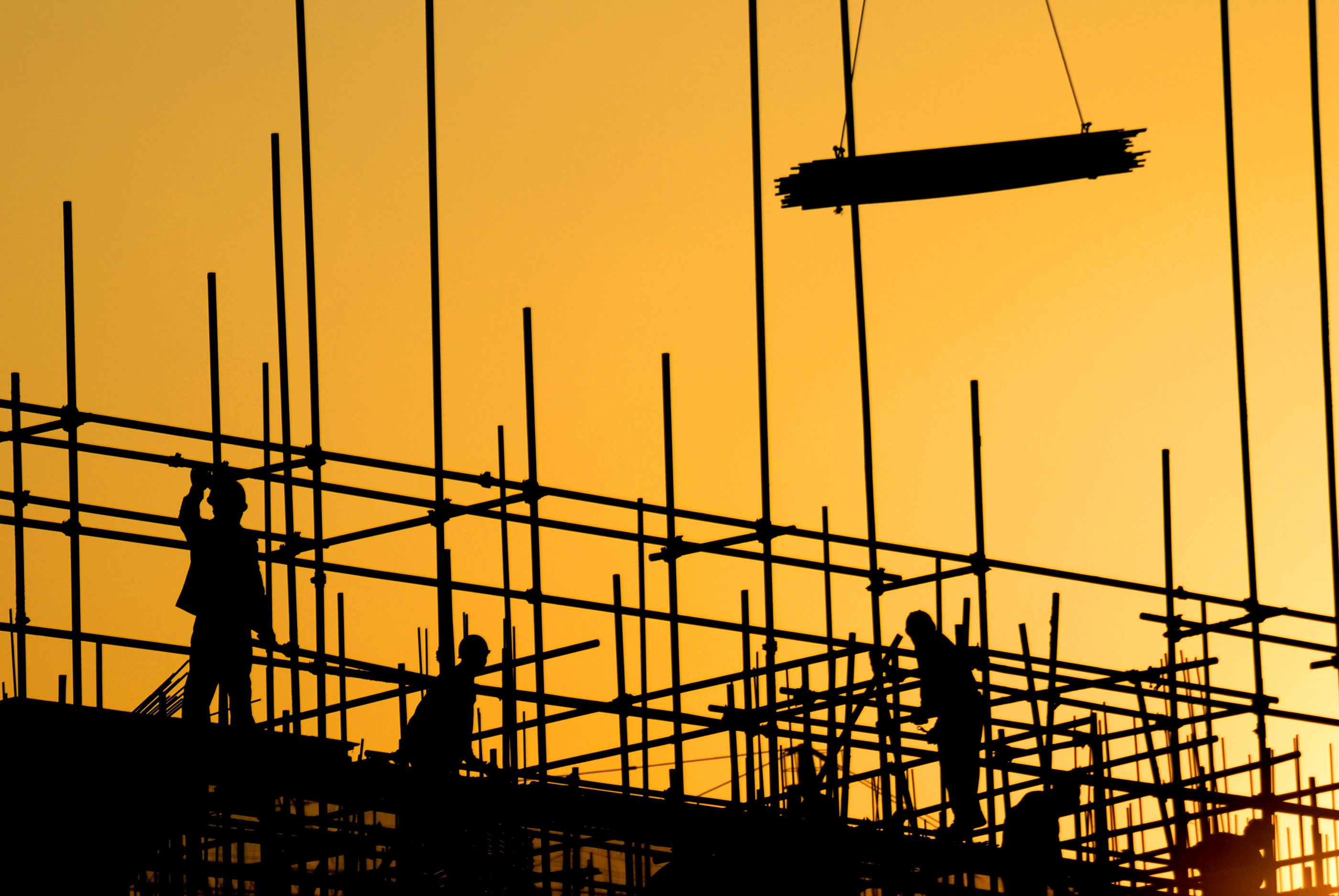 Construction workers on a scaffolded construction site, silhouetted against an evening sky.