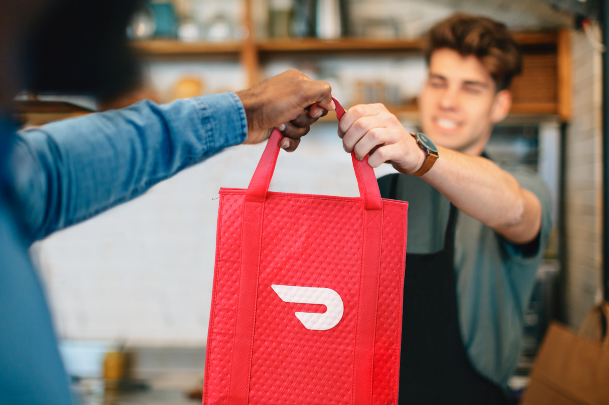 Man wearing an apron handing over a red bag with a DoorDash logo on it