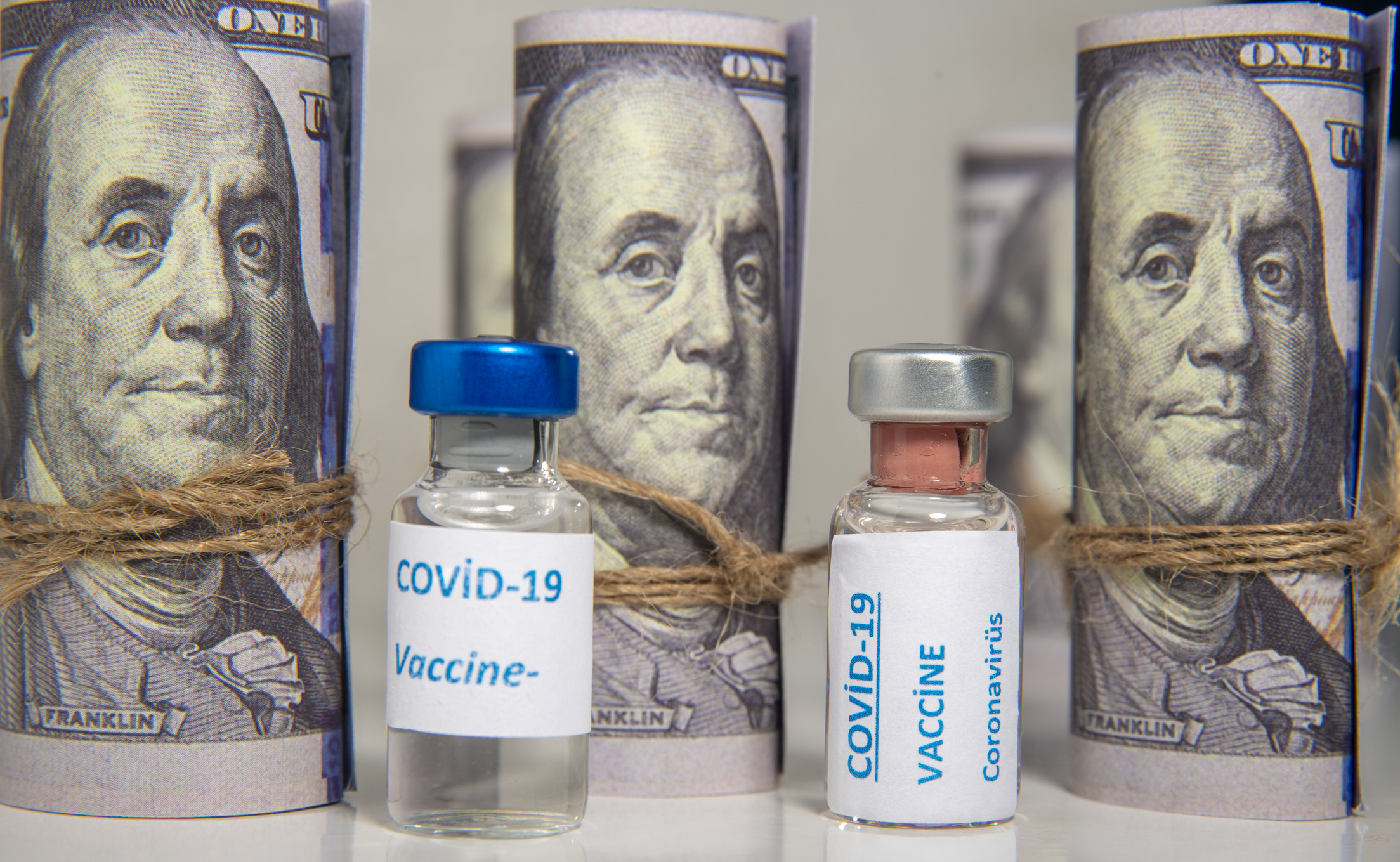 Vials labeled COVID-19 Vaccine, in front of a backdrop of rolled-up $100 bills