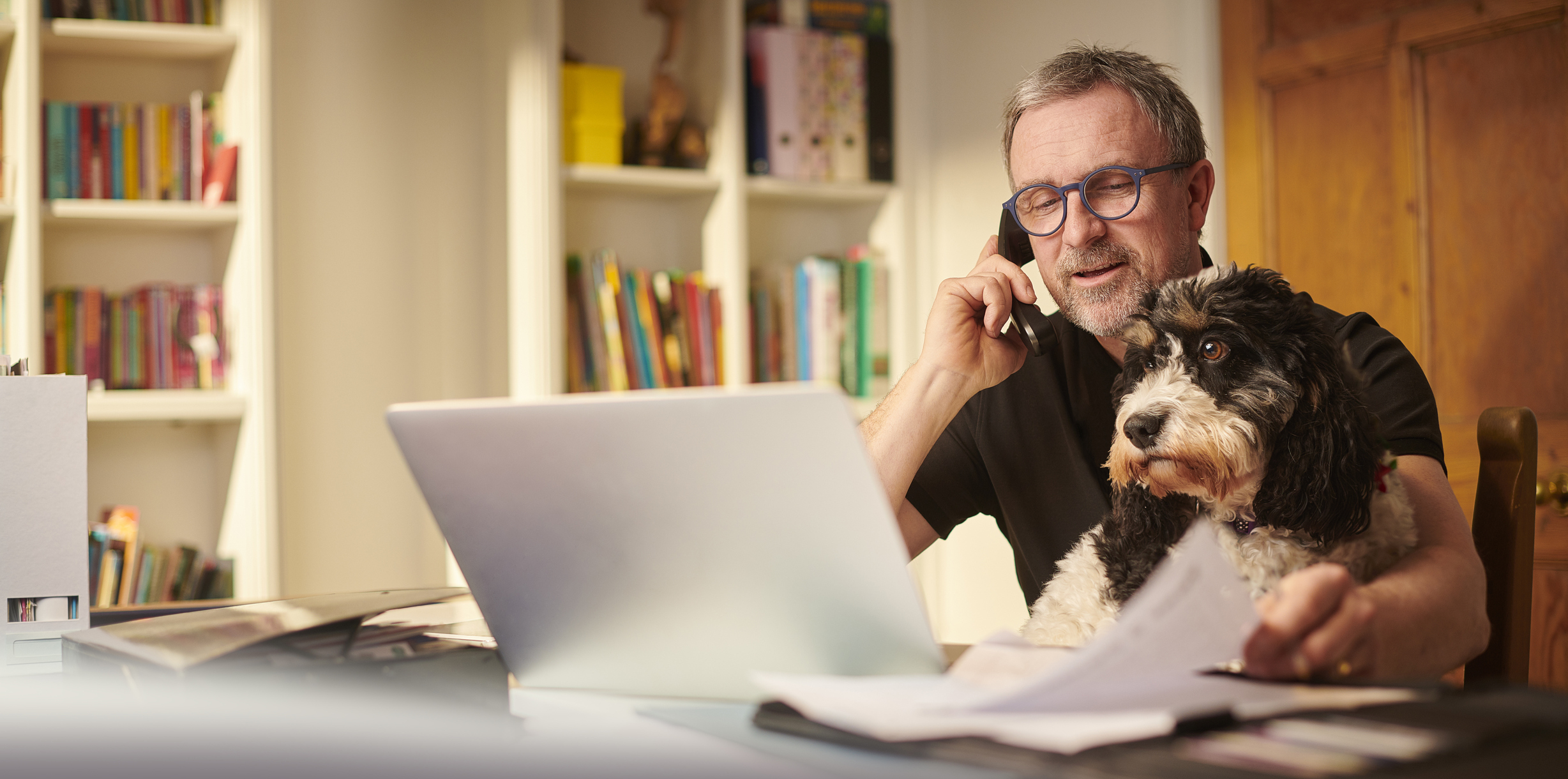 Older man at laptop holding phone to ear with dog on lap