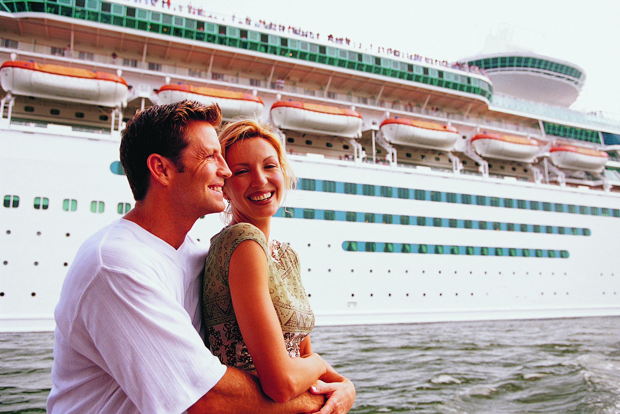 A couple poses in front of a cruise ship at port.
