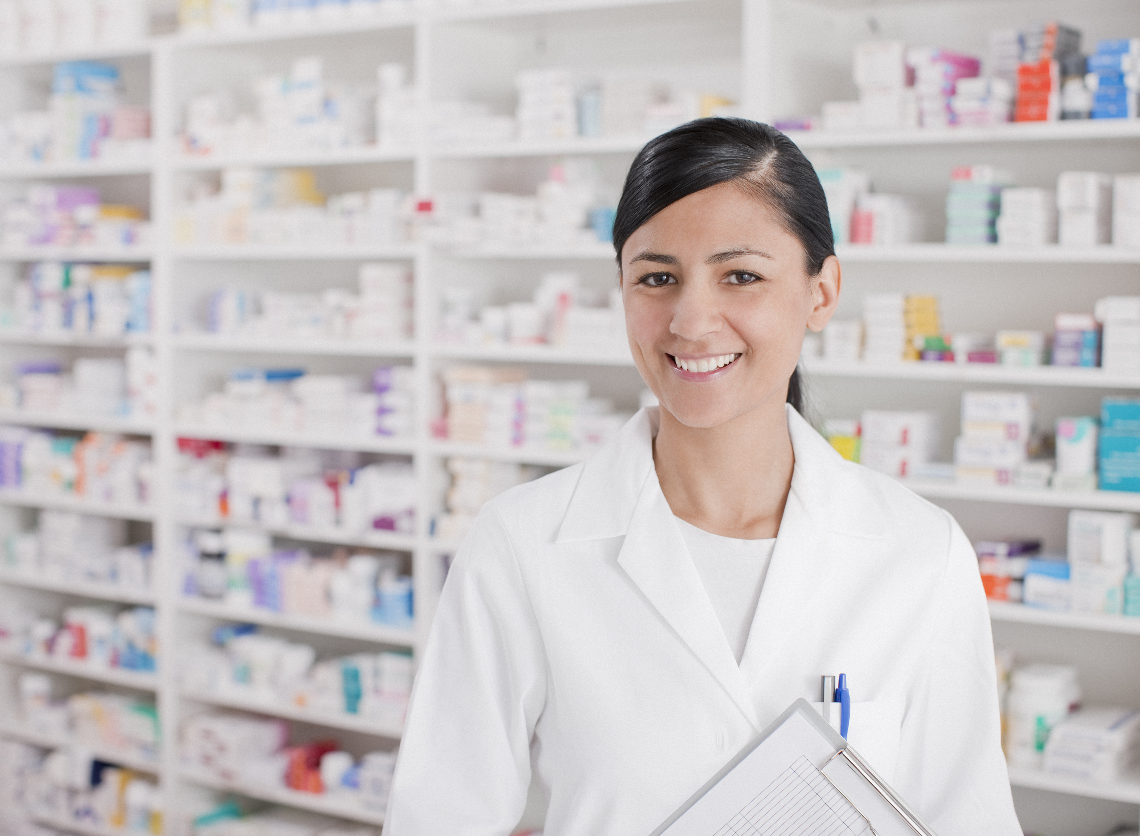 Smiling person in white lab coat, with shelves of pharmaceutical drugs behind.