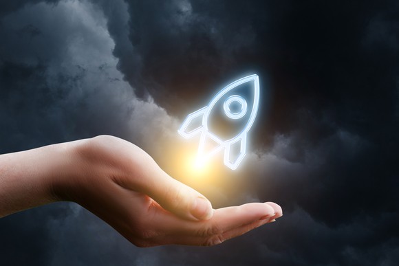 A rendering of a rocket ship blasts off from the palm of a hand.