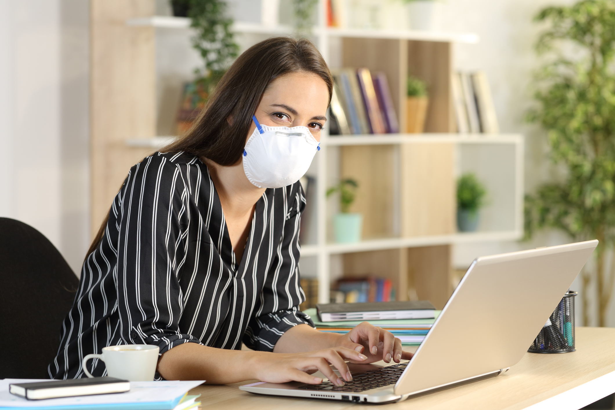 A young woman wearing a face mask works with a laptop in her living room.