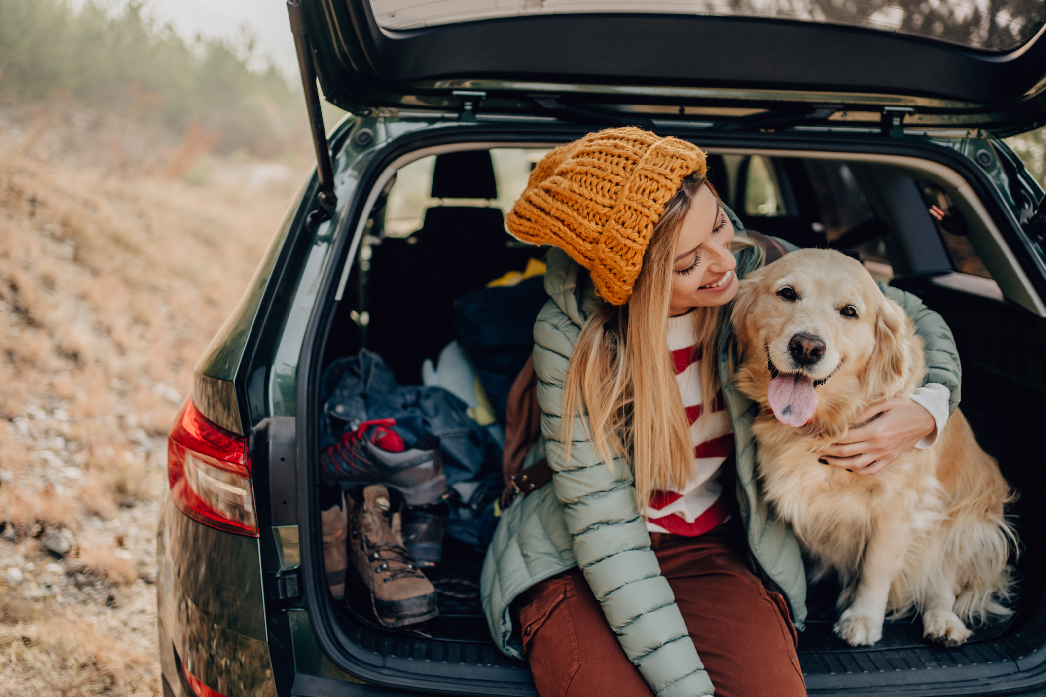 A young woman smiling sitting with her dog in the back of a car with a hatchback.