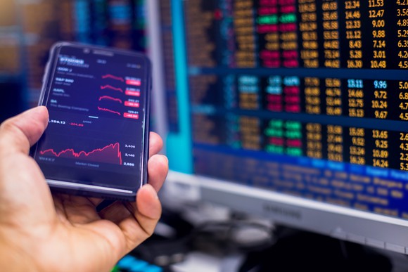 A smartphone with visible stock quotes being held next to a computer with real-time trade data.
