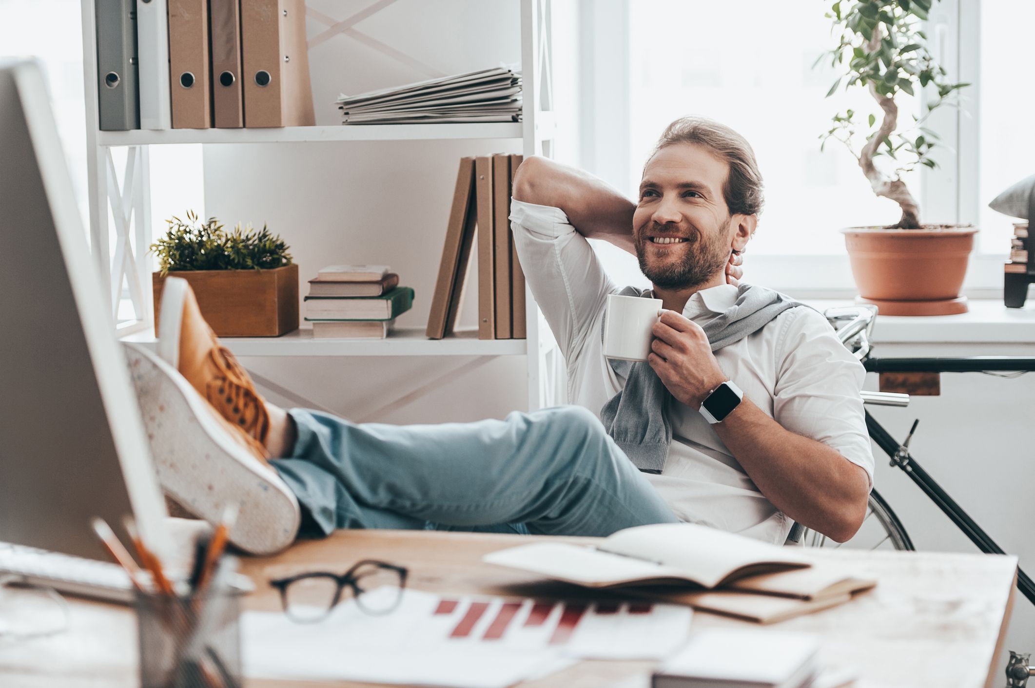 A relaxed man reclines with his feet on his desk while holding a cup of coffee.