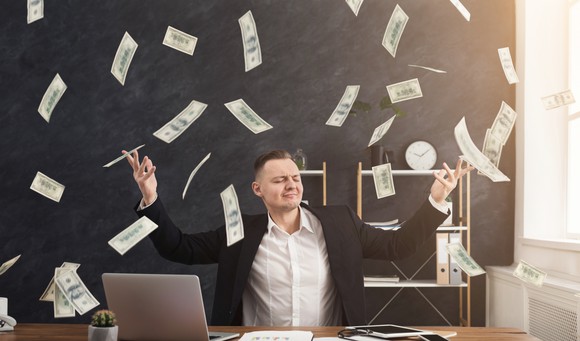 A man in a suit holds his hands up as money flies around the room.