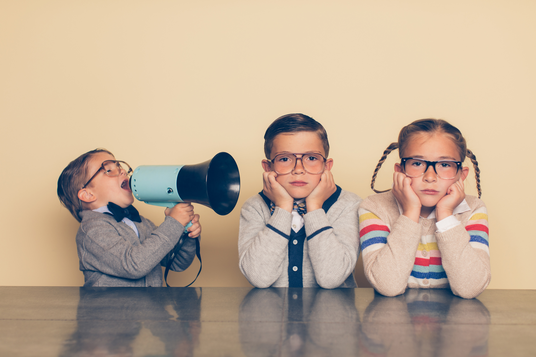 Two kids plug their fingers in their ears as a third kid shouts into a megaphone.