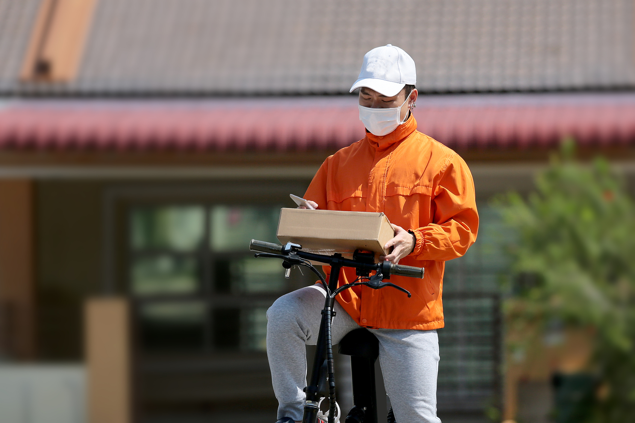 A deliveryman on a bike looks at a package in hishands wearing a mask.