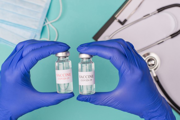 A healthcare professional's hands hold two vials labeled Vaccine: COVID-19.