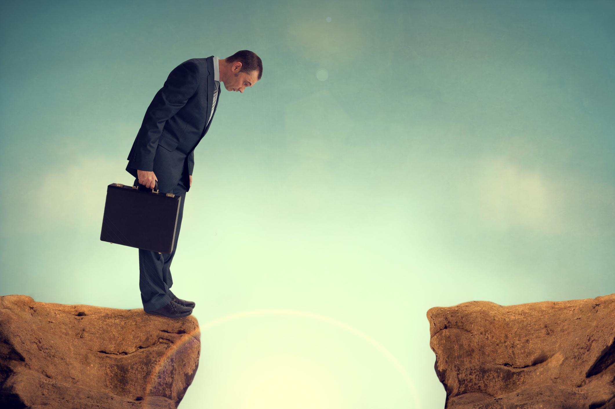A businessman peers over the edge of a cliff at his feet.