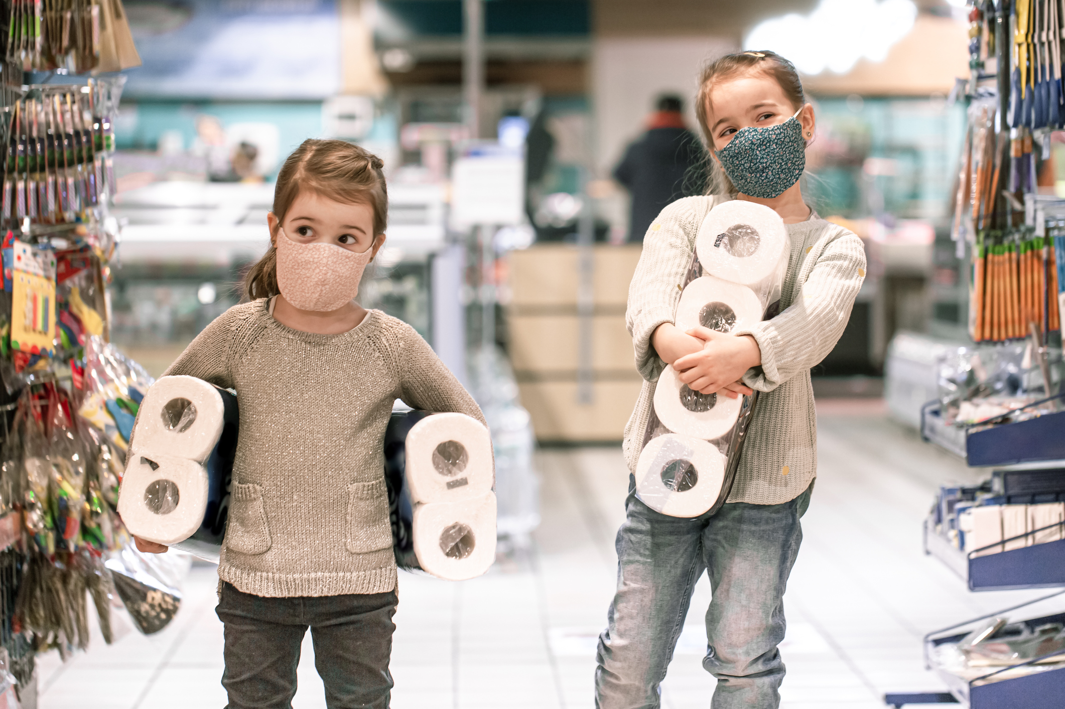 Two young children wearing face masks and carrying large packages of bathroom tissue in a retail store.