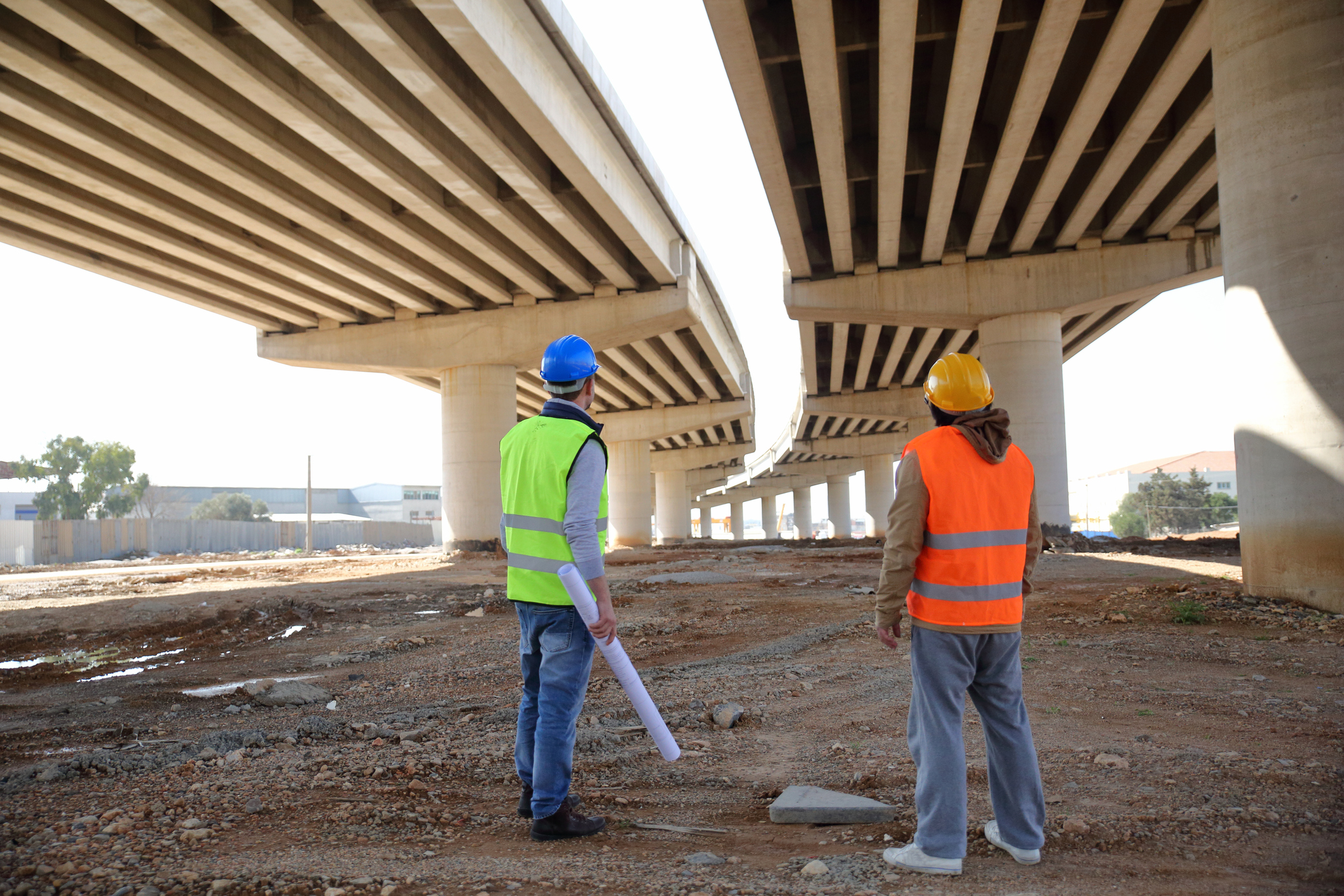 Two workers stand underneath a highway project.