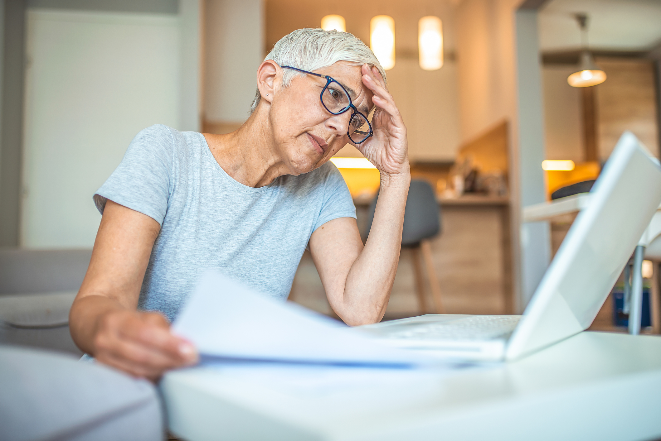 Older person at laptop holding head with one hand and document with another.