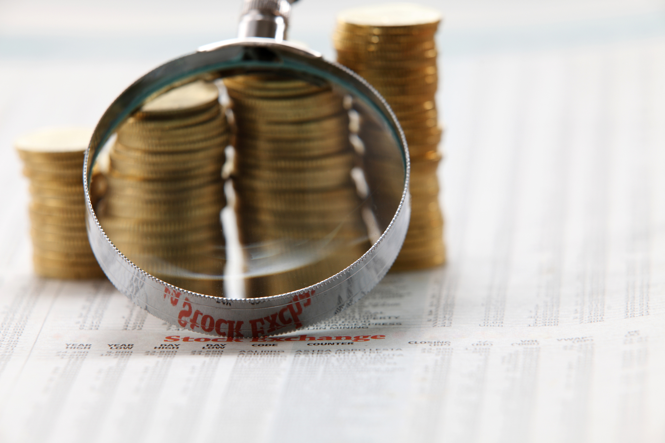 A magnifying glass being held above ascending stacks of coins that are on top of a financial newspaper.