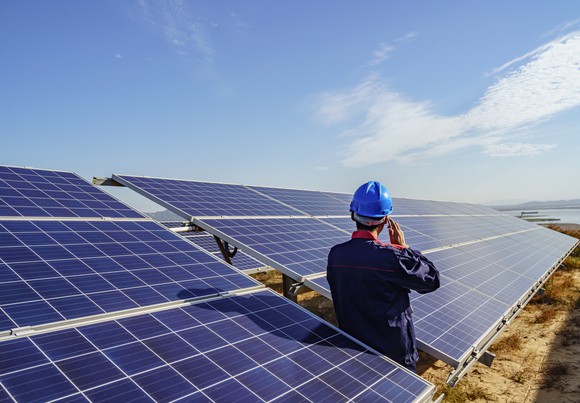 A worker leans against a solar panel while talking on his cell phone. 