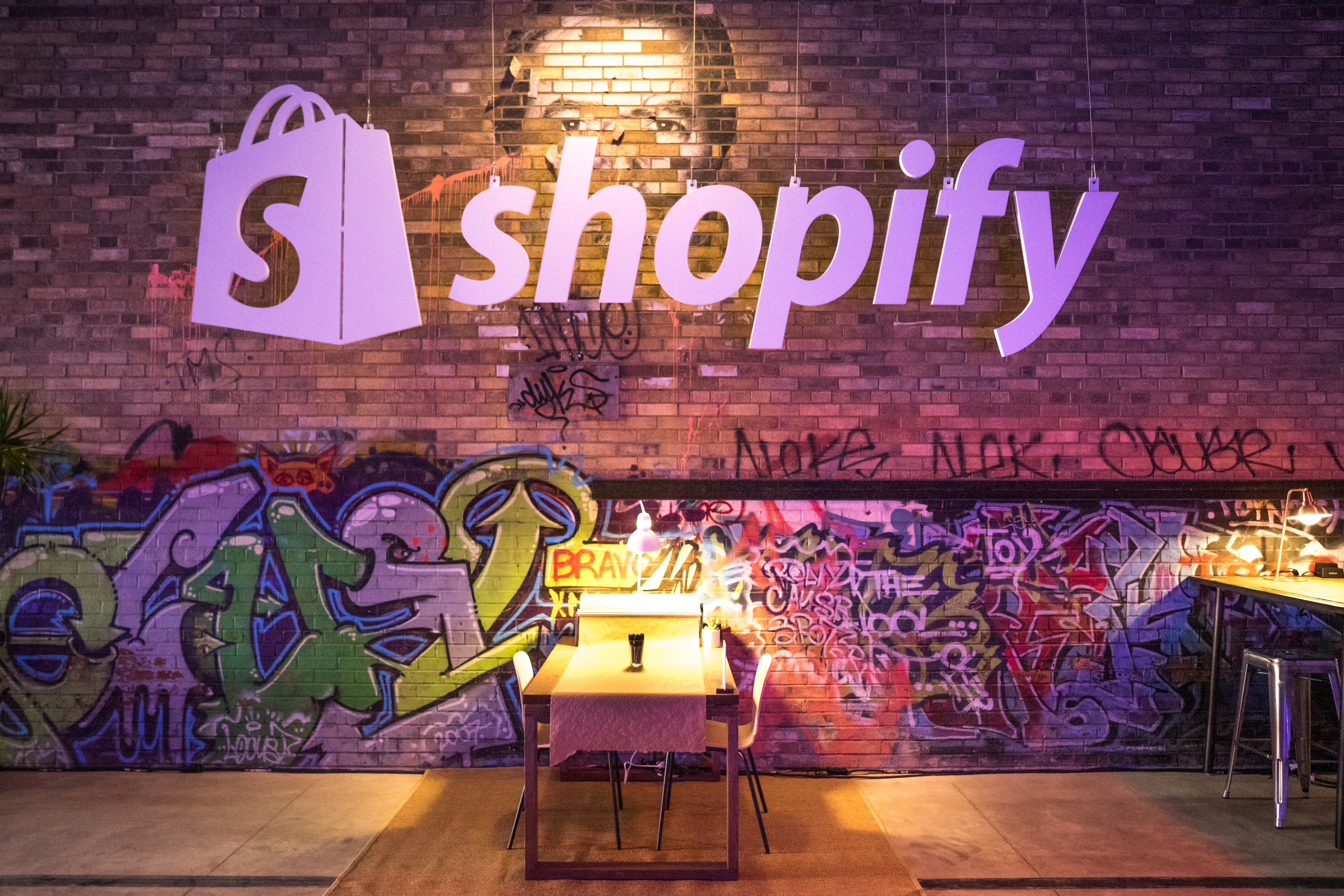 The Shopify logo above a cafeteria table and a brick wall decorated with graffiti.