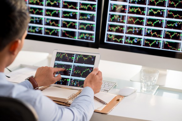 A stock trader analyzing dozens of charts on computer monitors and a tablet. 