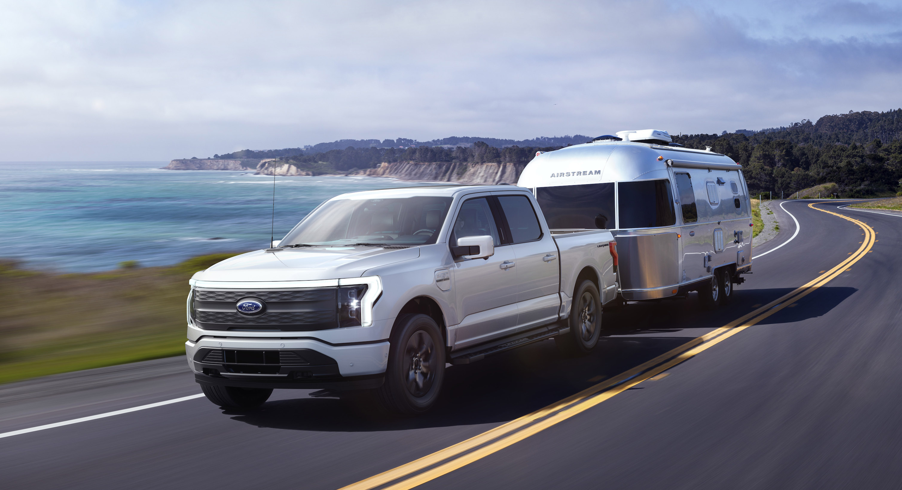 A silver 2022 Ford F-150 Lightning Lariat, an electric pickup truck, shown towing a camping trailer on a coastal road.