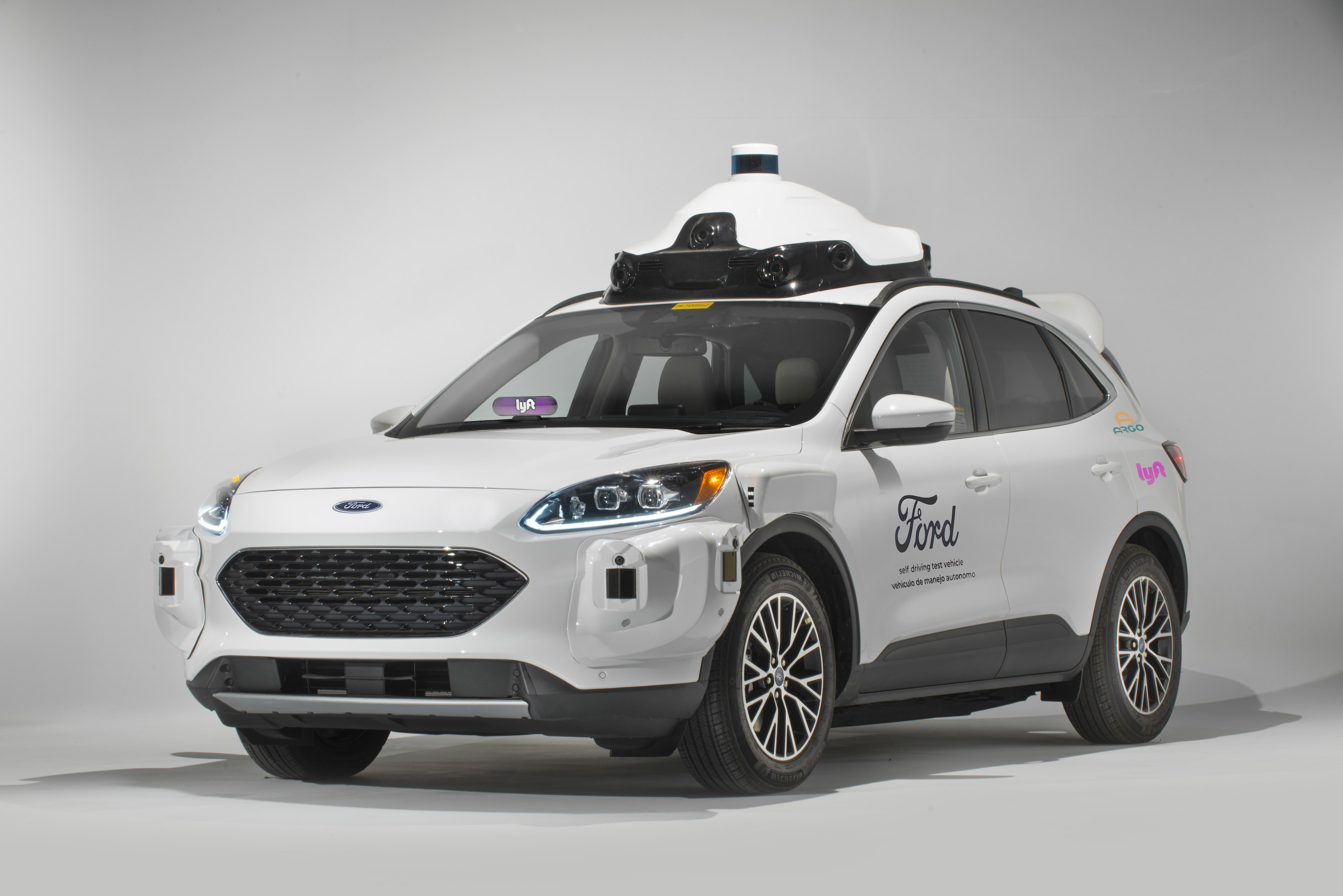 A white Ford Escape Hybrid SUV with visible self-driving sensor hardware and Ford, Lyft, and Argo AI logos.