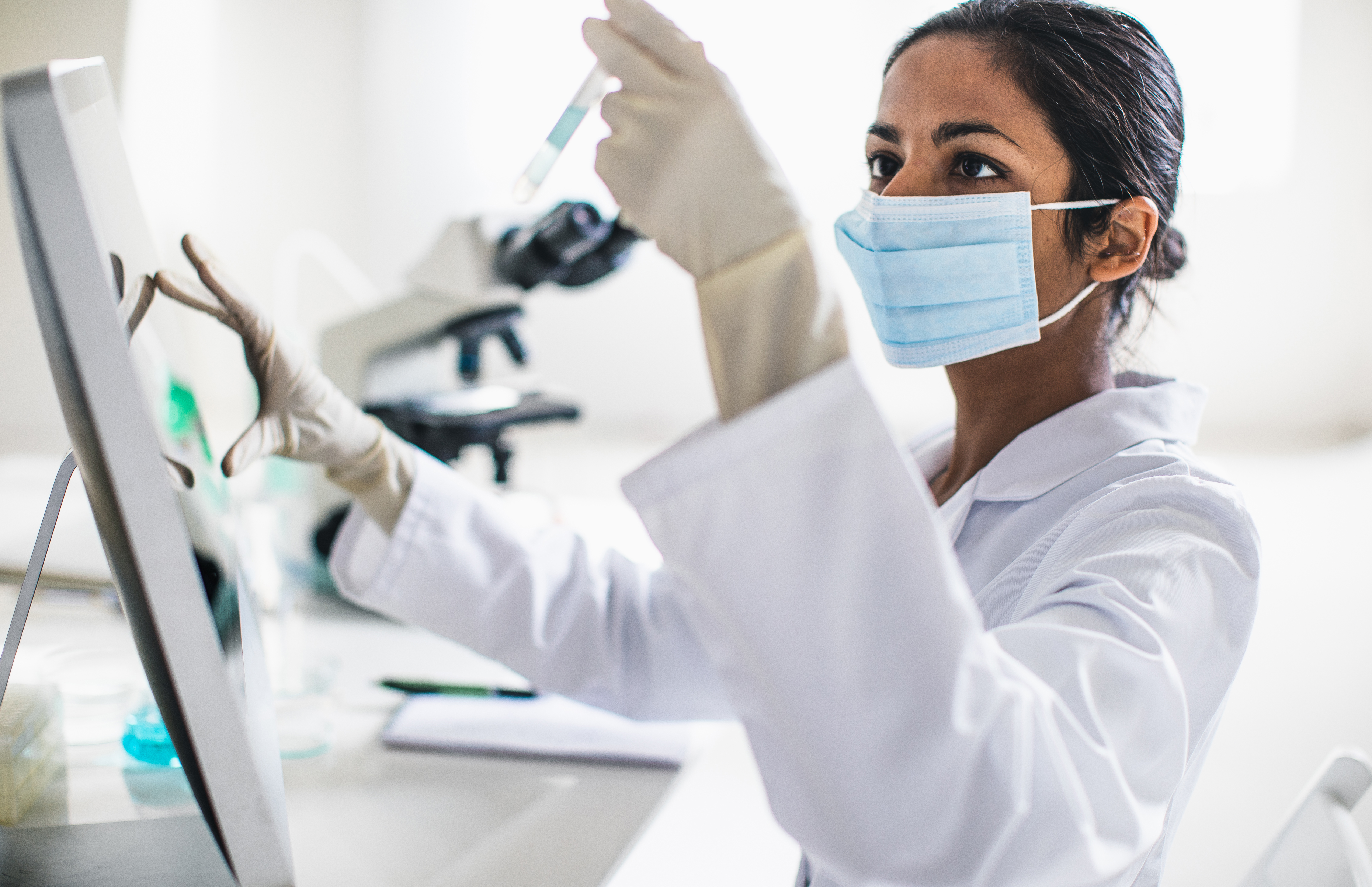 Woman with dark hair and skin. wearing a surgical mask and a white lab coat holds up a vial in one hand, with her other hand working on a touch-screen device in a laboratory setting.