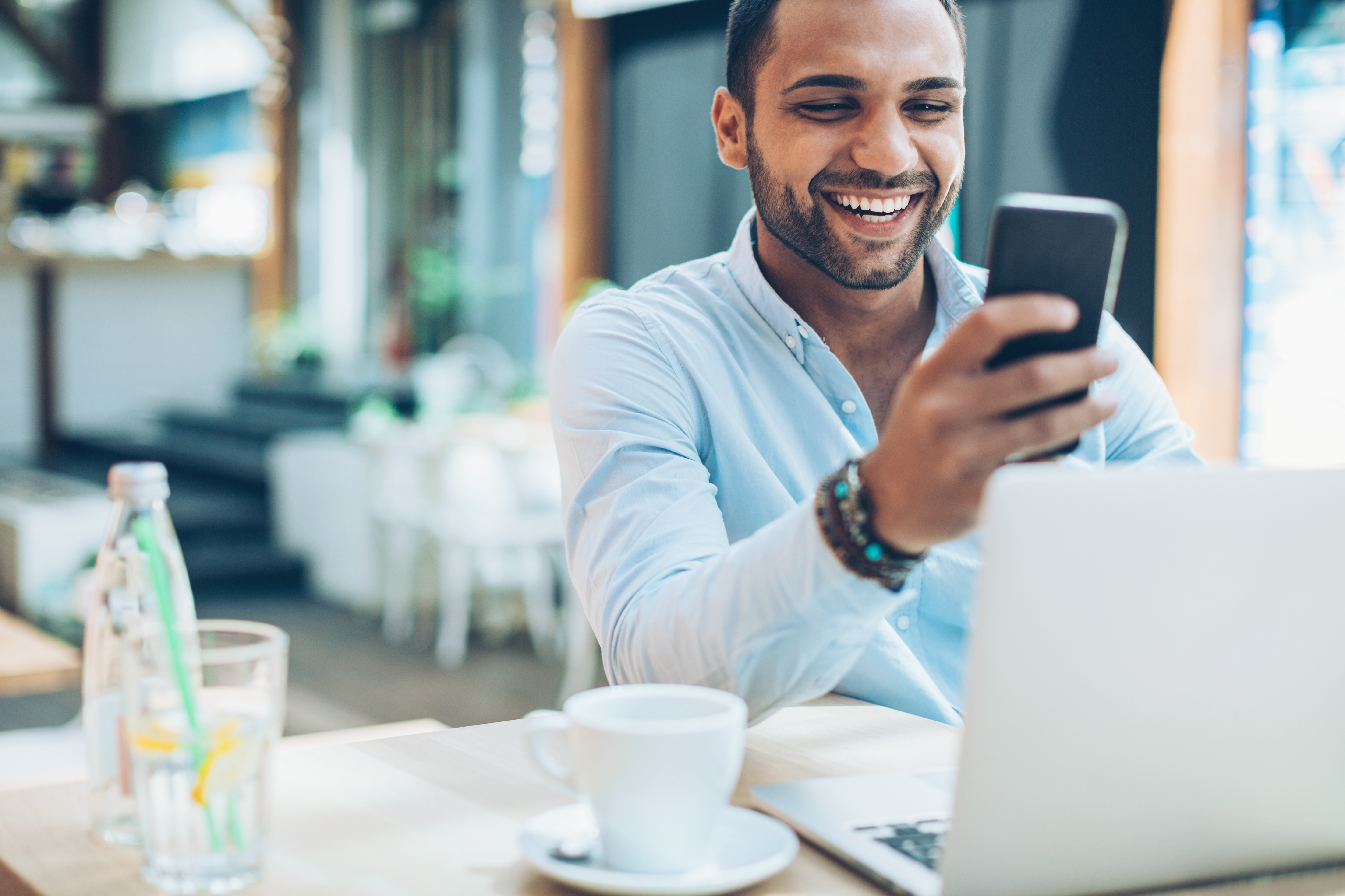 Man wearing light blue button-down shirt sits at a table with a laptop, glass of water, and mug nearby, smiling while looking at his smartphone.
