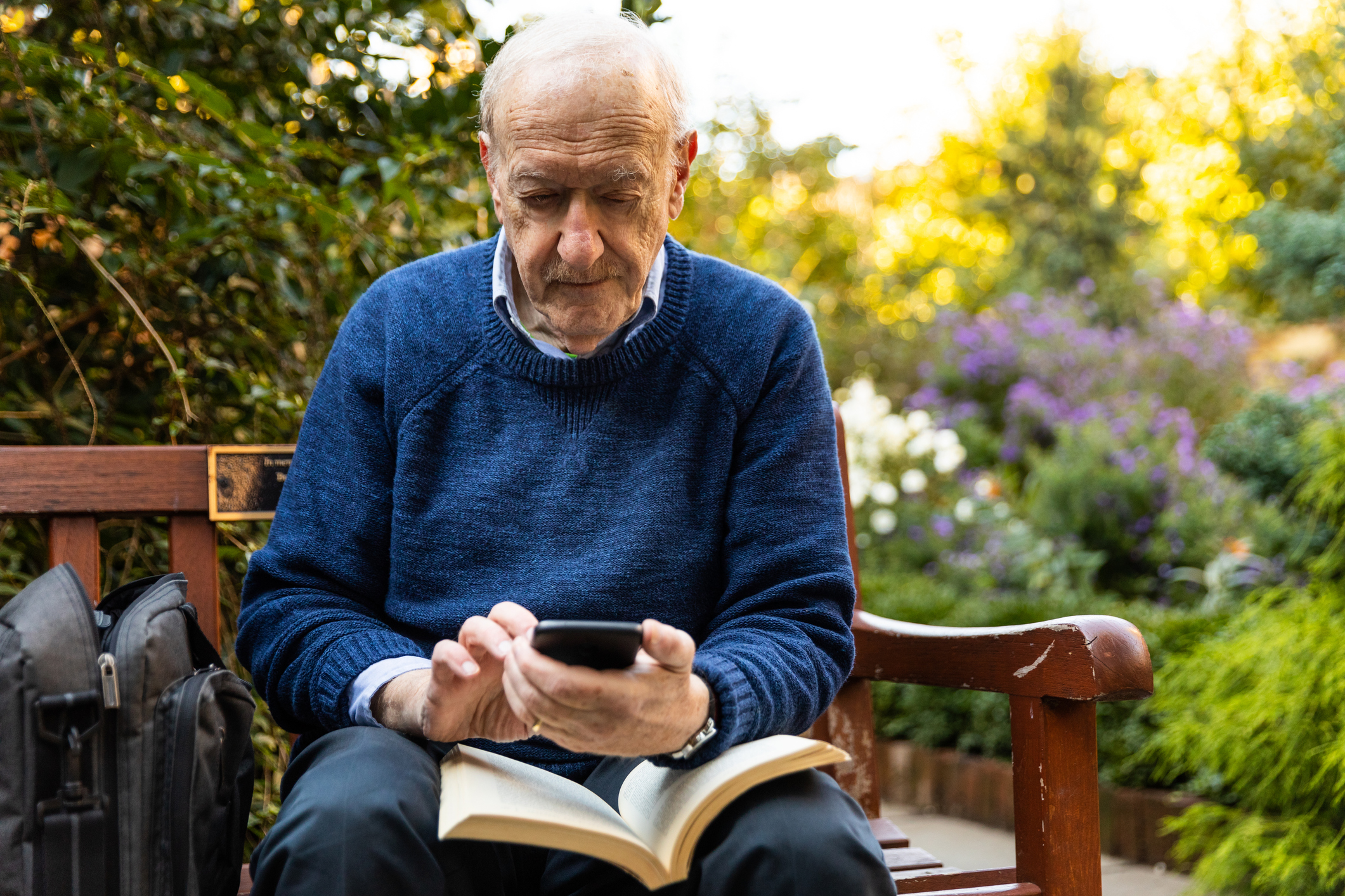 Older man sitting on bench with book in lap holding phone