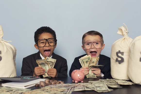 Two young boys in suits holding cash and surrounded by money bags.